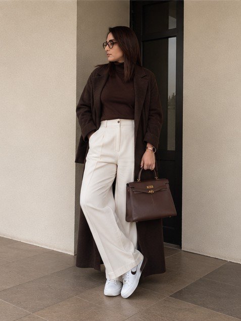 Farkhana Khan in a long brown coat and cream trousers, holding a brown handbag